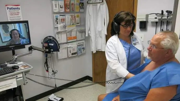 Physician with headset performing an auscultation on a patient while a remote doctor displayed on screen listens in.